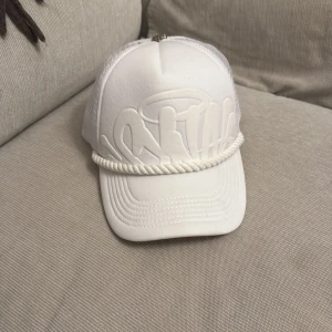 Vit keps med broderad text - Snygg vit keps med broderad text framtill och mesh-paneler på sidorna och baksidan. Kepsen har en dekorativ snodd framtill och justerbar passform. Perfekt för en avslappnad stil.