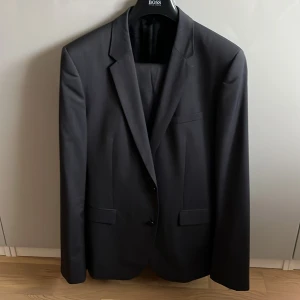 Svart kostym från Boss - Elegant svart kostym från Boss med kavaj och matchande byxor. Kavajen har två knappar och klassiska slag. Insidan av kavajen har stiliga röda detaljer. Perfekt för formella tillfällen. Modellen är specialbeställd och längre än normalt. Skulle säga att den passar fr 190 till 195 längd. Byxornas yttre ben längd är 112cm och inre längd 78cm. Om ni har fler funderingar är det bara att meddela.