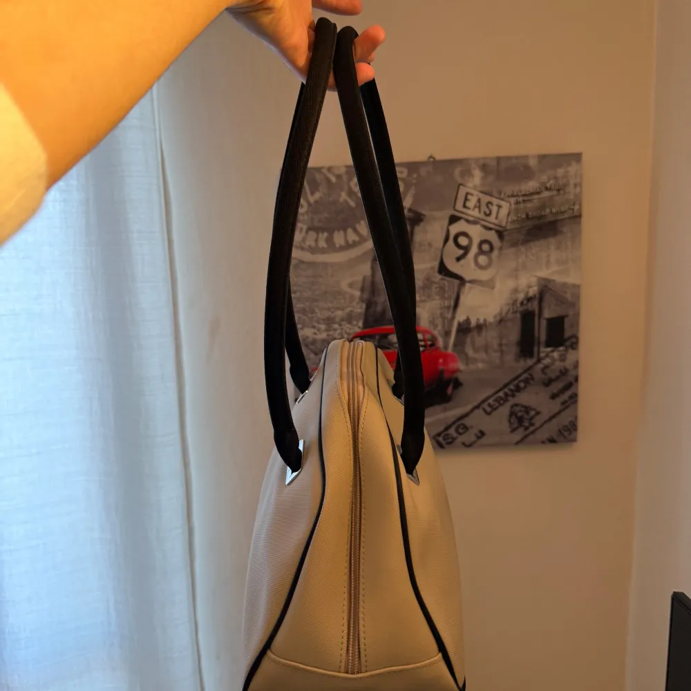 Snygg beige handväska från Lacoste med svarta handtag och detaljer. Väskan har en dragkedja upptill och är perfekt för att bära dina dagliga nödvändigheter. Den stilrena designen gör den lätt att matcha med olika outfits.. Laukut & Käsilaukut.