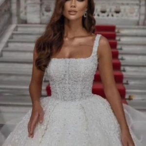 Vit bröllopsklänning med pärlor - Elegant vit bröllopsklänning med pärlor och ett vackert mönster. Klänningen har en klassisk lång design med axelbandslös stil och en voluminös kjol. Perfekt för en sagolik bröllopsdag.