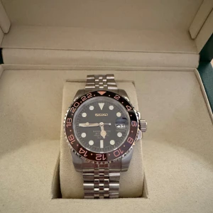 Seiko GMT Rootbeer - Säljer en stilren Seiko klocka med en svart urtavla och en röd-svart bezel. Klockan har en robust länkarmband i silverfärg och tydliga markeringar på urtavlan. Perfekt för den som söker en kombination av funktion och stil.