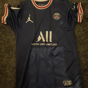 Blå PSG fotbollströja med Messi 30 - Snygg blå PSG-tröja från Jordan med Messi 30 på ryggen. Tröjan har korta ärmar och klubbens emblem på bröstet. Den är dekorerad med vita och röda detaljer vid krage och ärmslut. Perfekt för fotbollsfans!