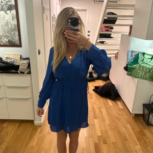 Blå klänning från H&M - Säljer en snygg blå klänning från H&M med långa ärmar och v-ringad design. Klänningen har en luftig passform och är perfekt för olika tillfällen. Den är tillverkad i ett lätt och bekvämt material.