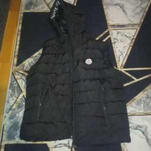 Svart dunväst från Moncler - Säljer en svart dunväst från Moncler med huva och dragkedja. Västen har ett quiltat mönster och två praktiska fickor med dragkedja framtill. Perfekt för kyligare dagar.