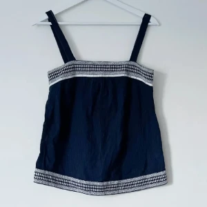 Mörkblå linnetopp med vita detaljer - Snygg mörkblå linnetopp med breda axelband och vita mönstrade detaljer vid byst och nederkant. Perfekt för en somrig look. Storlek XS
