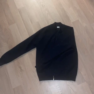 Svart hoodie med dragkedja - Säljer en stilren svart hoodie med dragkedja. Perfekt för en avslappnad look. Den har en normal passform och är tillverkad i ett mjukt material för extra komfort.