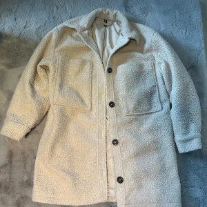 Beige teddyjacka med knappar - Mysig beige teddyjacka från H&M med knappar framtill och stora fickor. Perfekt för kyliga dagar med sin mjuka och varma känsla. Jackan har en klassisk krage och långa ärmar med knappdetaljer vid ärmsluten.