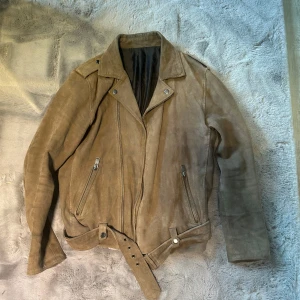 Beige mockajacka  - Snygg beige mockajacka med dragkedja och bälte i midjan. Jackan har långa ärmar och en klassisk bikerstil med snedställd dragkedja och två fickor med dragkedjor framtill.