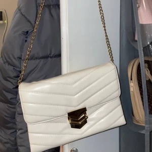Vit axelväska med kedja - Snygg vit axelväska med quiltad design och guldfärgad kedja. Väskan har en elegant låsdetalj i guld och är perfekt för att ge en stilren touch till din outfit.