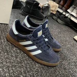 Adidas Spezial sneakers i blå mocka - Snygga Adidas Spezial sneakers i blå mocka med klassiska vita ränder och brun sula. Perfekta för en stilren och sportig look. Skorna har snörning och en diskret gulddetalj på sidan.