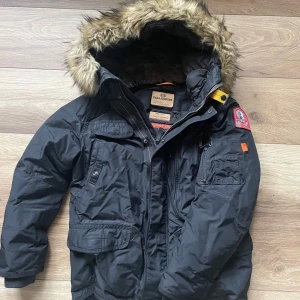 Svart parajumper jacka med pälsdetalj - Säljer en svart jacka från Parajumpers med avtagbar huva och pälsdetalj. Jackan har flera fickor och en dragkedja framtill. Perfekt för kalla dagar med sin varma och skyddande design. Ny skick