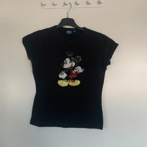 Svart Disney t-shirt med paljetter - Säljer en svart t-shirt från Disney med en paljettdekoration av en känd muskaraktär på framsidan. T-shirten har korta ärmar och en normal passform. Perfekt för dig som älskar klassiska tecknade figurer!