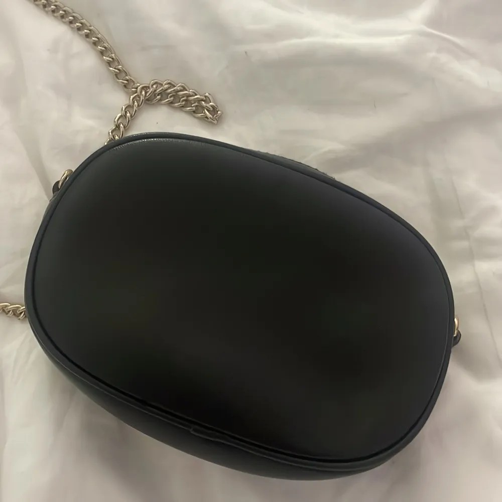Snygg svart axelväska med guldfärgad kedja och dekorativt spänne på framsidan. Väskan har en dragkedja upptill och e från H&M. Väskan har en liten ficka i sig och är väldigt fin o lätt att styla.. Laukut & Käsilaukut.