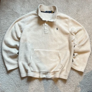 Ralph Lauren Beige fleece tröja  - Säljer en mysig beige fleece tröja från Polo Ralph Lauren. Tröjan har en knappslå framtill och en praktisk känguruficka. Perfekt för kyliga dagar med sin mjuka och varma känsla. Nypris ca 2000kr