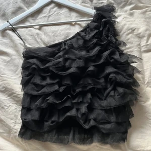 Svart volangig oneshoulder topp Stockholm LM - Snygg svart oneshoulder topp med volanger. Perfekt för en kväll ute eller en speciell tillställning. Den unika designen med volanger ger en trendig och elegant look.