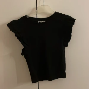 Svart topp med volangärmar från Zara - Säljer en stilren svart topp från Zara med volangdetaljer på ärmarna. Perfekt för att ge en extra touch till din outfit. Toppen är i en mjuk och bekväm kvalitet.