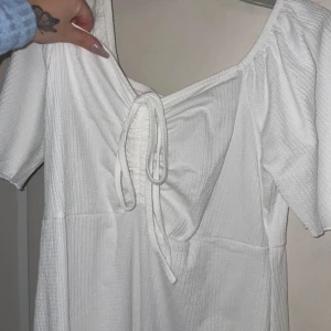 Vit klänning med knytning - Säljer en söt vit klänning med knytning framtill och korta ärmar. Perfekt för sommarens varma dagar. Klänningen har en v-ringad design och är gjord i ett lätt och luftigt material.