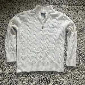Ralph Lauren Knitted Half Zip - Säljer en stilren vit stickad tröja från Polo Ralph Lauren med kabelstickat mönster och en halv dragkedja vid halsen. Tröjan har långa ärmar och ribbade muddar för en klassisk look. Perfekt för kyligare dagar! 