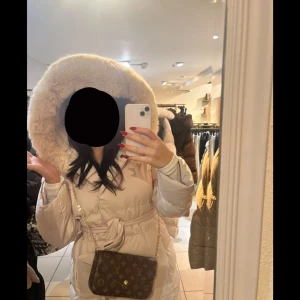 Vit dunjacka med pälsdetaljer - Snygg och varm vit dunjacka med stor pälskantad huva. Jackan har ett bälte i midjan för en figursydd look och långa ärmar. Perfekt för kalla dagar när du vill hålla stilen samt aldrig använd, köpt men kunde ej lämnas tillbaka 