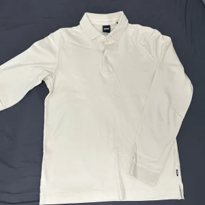 Vit långärmad pikétröja från Boss - Säljer en stilren vit långärmad pikétröja från Boss. Tröjan har en klassisk krage och knappar framtill. Perfekt för en avslappnad men ändå elegant look. Passar bra till både jeans och chinos.