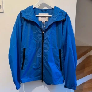 Blå vindjacka från Woolrich - Säljer en snygg blå vindjacka från Woolrich. Jackan har en praktisk dragkedja framtill och två fickor med dragkedjor. Perfekt för blåsiga dagar med sin lätta och bekväma design. 🌀