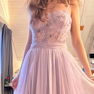 Ljuslila balklänning med blommig broderi - Superfin balklänning i ljuslila med en korsettinspirerad överdel och med ett vackert blommigt broderi. Den har en härligt luftig kjol och markerad midja som ger en jättefin siluett. Använd bara en gång, så den är i toppskick! 