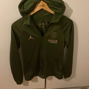 Grön huvtröja från Jordan x PSG - Säljer en grön huvtröja från Jordan i samarbete med Paris Saint-Germain. Tröjan har en dragkedja framtill och är dekorerad med PSG-loggan och Qatar Airways-tryck. Perfekt för sportiga tillfällen eller som en stilren vardagslook.