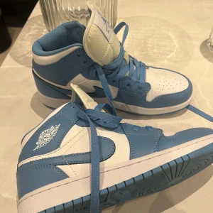 Nike Air Jordan 1 Mid blå/vit - Snygga Nike Air Jordan 1 Mid sneakers i blått och vitt skinn. Klassisk siluett med perforerad tå, blå snörning och ikoniska Air Jordan-loggan på sidan. Perfekta för dig som vill sticka ut med stilrena och sportiga vibes.