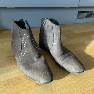 Bruna mocka boots med nitar - Snygga bruna boots i mocka med dekorativa nitar och dragkedja på sidan. Perfekta för en stilren look med en spetsig tå och låg klack.