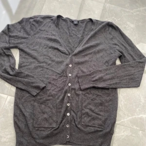 Grå kofta från Amisu - Säljer en stilren grå kofta från Amisu. Den har en v-ringad design med knappar framtill och praktiska fickor. Perfekt för lager-på-lager under kyligare dagar.