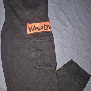 Svarta byxor med orange detalj - Snygga svarta byxor med en cool orange detalj och texten 'whatever' på benet. Byxorna har en ficka på sidan och är perfekta för en avslappnad stil.