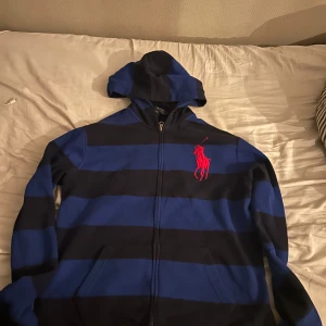 Randig hoodie från Ralph Lauren - Säljer en snygg randig hoodie från Ralph Lauren i blått och svart. Den har en dragkedja framtill och en stor röd logga på bröstet. Perfekt för en avslappnad stil.