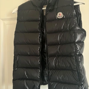 Svart dunväst från Moncler - Snygg svart dunväst från Moncler med glansig yta och klassisk logga på bröstet. Perfekt för kyligare dagar när du vill hålla stilen. Västen har en dragkedja framtill och är lätt att matcha med olika outfits.