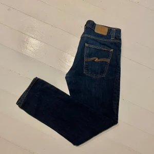 Mörkblå jeans från Nudie Jeans - Säljer ett par mörkblå jeans från Nudie Jeans med klassisk femficksdesign och orange sömmar. Jeansen har en rak passform och är tillverkade i slitstarkt denim. Perfekta för en avslappnad stil. Byxorna är i väldigt bra skick och knappt använda. Hör av dig om du har några funderingar!