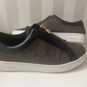 Bruna sneakers från Michael Kors - Säljer dessa snygga bruna sneakers från Michael Kors med svart läderdetalj och guldfärgad dragkedja för 1000kr då jag köpte de för 1500kr och eftersom dem inte kommit till användning då de bara använts ett par gånger. Dessa är i storlek 37. 