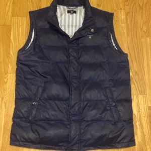 Mörkblå Gant väst - Säljer en stilren mörkblå dunväst från Gant. Västen har en dragkedja framtill och två praktiska fickor med knappar. Insidan är fodrad med ett randigt mönster. Har en liten defekt vid ryggen men syns knappt 