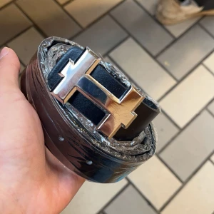 Svart bälte med silverfärgat spänne -  Priset kan diskuteras vid snabb affär , Snyggt svart bälte med ett iögonfallande silverfärgat spänne i form av ett H. Perfekt för att ge en stilren touch till din outfit. Bältet är tillverkat i ett slätt material och har flera justeringshål för bästa passform.