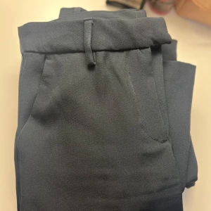 Svarta kostymbyxor  - Snygga svarta kostymbyxor med hög midja och bälteshällor. Byxorna har en klassisk rak passform, diskreta fickor både fram och bak och slits ner till. Perfekta för en stilren look.