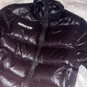Svart pufferjacka från NOCTA - Snygg svart pufferjacka från NOCTA med Nike-logga. Jackan har en hög krage och dragkedja framtill. Perfekt för kyliga dagar med sin stilrena design och glansiga yta.