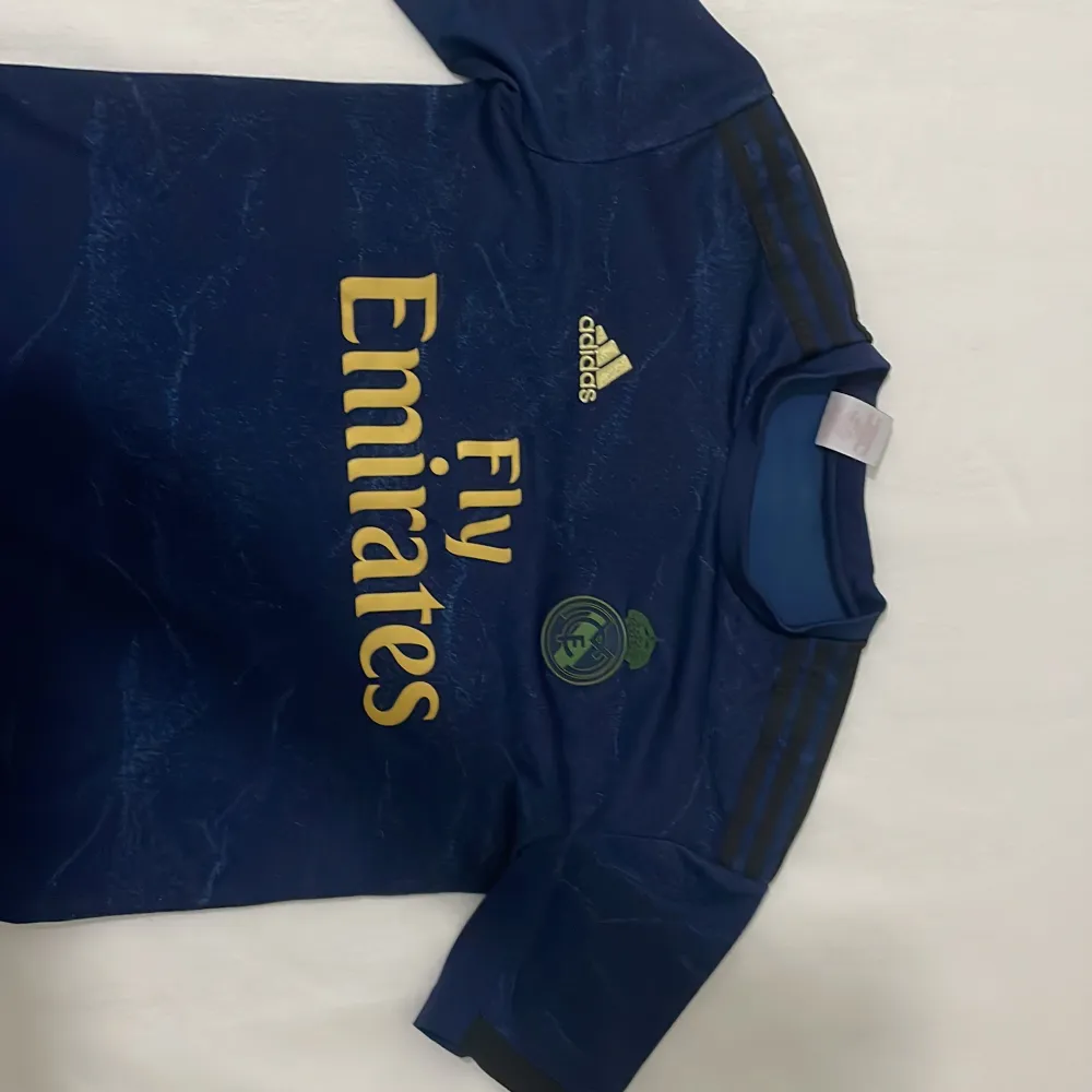 Snygg blå Real Madrid fotbollströja från Adidas med Fly Emirates-logga i guld. Tröjan har korta ärmar och ett subtilt mönster över hela tyget. Perfekt för alla fotbollsfans!. T-paidat.