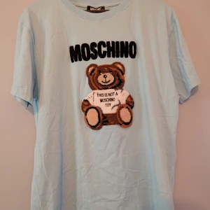 Ljusblå t-shirt med nalle från Moschino - Säljer en ljusblå t-shirt från Moschino med en stor nalleapplikation på framsidan. Texten 'This is not a Moschino toy' är broderad på nallens tröja. Perfekt för dig som gillar lekfull design!