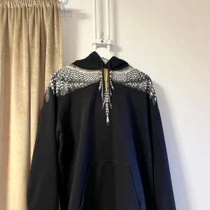 Svart hoodie från Marcelo Burlon - Snygg svart hoodie från Marcelo Burlon med ett unikt mönster i vitt, blått och rött över axlarna och ryggen. Hoodien har en stor ficka framtill och justerbar huva med snörning. Perfekt för en cool och avslappnad stil.