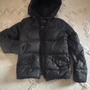Svart dunjacka från GANT - Snygg svart dunjacka från GANT med huva och dragkedja. Jackan har två praktiska fickor framtill och är perfekt för kyligare väder. Den är stilren och funktionell, vilket gör den till ett bra val för vintern.