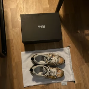 Versace Jeans Couture sneakers - Snygga sneakers från Versace Jeans Couture med ett unikt mönster i vitt och guld. Skorna har snörning och en robust sula. Perfekta för att ge din outfit en lyxig touch. Helt äkta såklart köpt på best secret kan sänka pris 