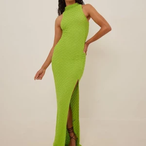 Grön långklänning med slits L oanvänd  - Snygg grön långklänning med hög hals och slits framtill. Klänningen har en texturerad yta och är ärmlös, vilket ger en elegant och modern look.