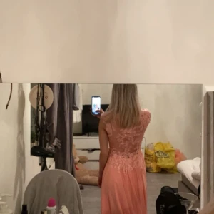 Persikofärgad långklänning med spetsdetaljer - Elegant persikofärgad långklänning med v-ringad spetsöverdel och ärmlös design. Perfekt för speciella tillfällen.