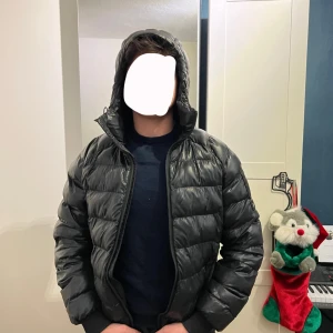 Svart dunjacka från Peak Performance - Snygg svart dunjacka från Peak Performance med huva och dragkedja. Jackan har en broderad logga på ärmen och är perfekt för kyliga dagar. Den är långärmad och har en stilren design. Dragkedjan behövs bytas ut därav priset! Modelen är 180 och väger 75kg.