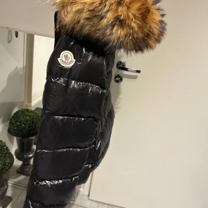 Svart dunjacka med pälsdetalj från Moncler - Snygg svart dunjacka från Moncler med en lyxig pälsdetalj på huvan. Jackan har en glansig finish och stängs med dragkedja. Perfekt för kalla vinterdagar. ( Storlek 3) 