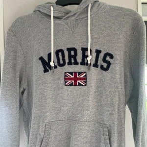 Grå hoodie från Morris - Säljer en grå hoodie från Morris med en broderad logga och brittisk flagga på bröstet. Tröjan har en justerbar huva med vita snören och en praktisk magficka. Perfekt för en avslappnad stil.
