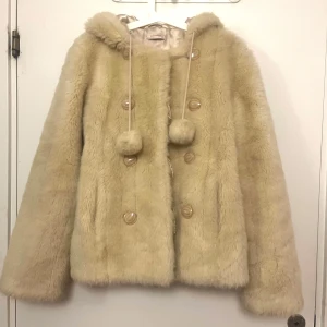 Beige pälsjacka med huva - Mysig beige pälsjacka med stora knappar och huva. Jackan har dekorativa bollar på snören framtill och en skön, fluffig känsla. Säljer för den är sitter stor på mig men annars är den sötaste jackan! På sista bilden kan man se att snoddarna som man stänger jackan med är lite förstörda men man kan stänga jackan fortfarande som vanligt dessutom så syns inte snoddarna när man stänger jackan. Står ingen storlek på jackan men jag skulle säga att den sitter som L, jag har storlek M